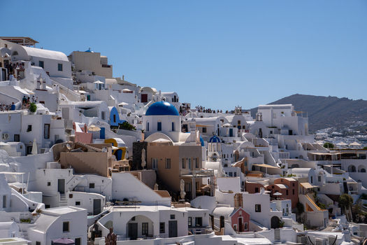 View of the iconic white houses and blue domed churches on the hillside with people walking along the narrow paths under a clear blue sky in Fira Greece. View of the iconic white houses and blue domed churches on the hillside with people walking along the narrow paths under a clear blue sky in Fira Greece.