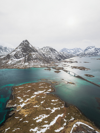 Aerial view of the Flakstadbruene bridge stretching across the shimmering turquoise waters amidst the rugged snow dusted landscape Flakstadbruene Nordland Norway. Aerial view of the Flakstadbruene bridge stretching across the shimmering turquoise waters amidst the rugged snow dusted landscape Flakstadbruene Nordland Norway.