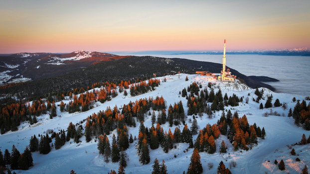 Aerial view of Col de la Faucille mountain pass featuring a communication tower snow covered slopes and pine trees under a golden sunset sky Gex Auvergne Rhone Alpes France. Aerial view of Col de la Faucille mountain pass featuring a communication tower snow covered slopes and pine trees under a golden sunset sky Gex Auvergne Rhone Alpes France.