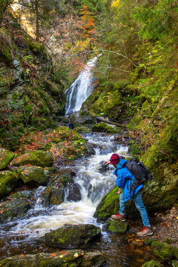 View of a hiker standing by a rushing stream with a waterfall in the background surrounded by mossy rocks and autumn foliage in Breitnau Baden Wurttemberg Germany. View of a hiker standing by a rushing stream with a waterfall in the background surrounded by mossy rocks and autumn foliage in Breitnau Baden Wurttemberg Germany.