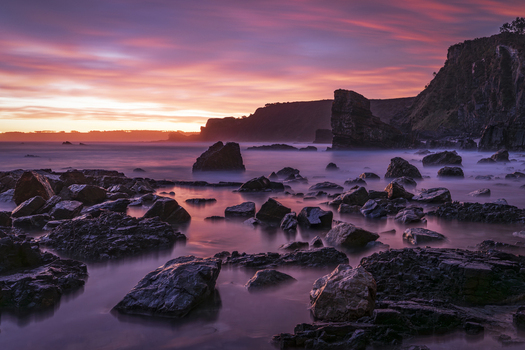 View of Playa de Portizuelo with jagged rocks and misty water under a vibrant purple sunset sky in Spain. View of Playa de Portizuelo with jagged rocks and misty water under a vibrant purple sunset sky in Spain.