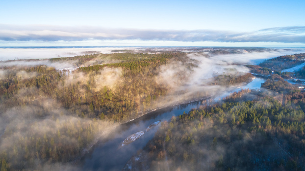 Aerial view of a winding river flowing through a dense forest blanketed in morning mist during a cold winter day in Latvia. Aerial view of a winding river flowing through a dense forest blanketed in morning mist during a cold winter day in Latvia.