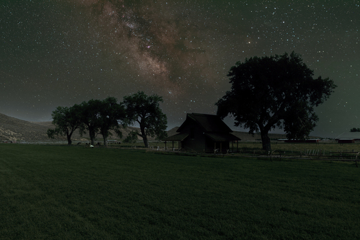 View of a historic 1800s ranch house set in a dark field under the Milky Way galaxy with mature trees in the foreground in Picabo Idaho United States.
