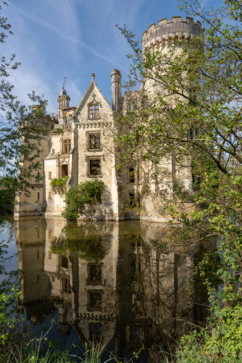 Les Trois Moutiers France 13 April 2024 View of the ruined Chateau de la Mothe Chandeniers reflected in its moat surrounded by lush green trees under a clear blue sky.