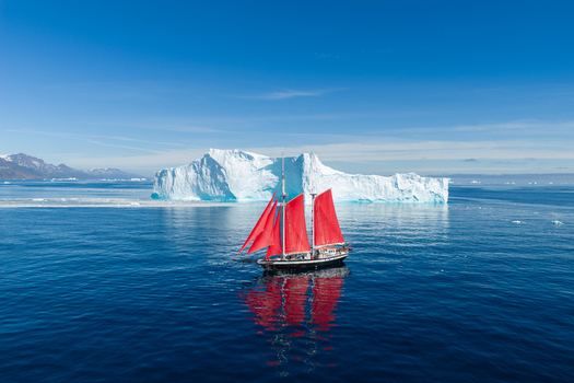 Aerial view of a sailboat with vibrant red sails navigating the deep blue waters near a massive white iceberg under a clear sky in Greenland. Aerial view of a sailboat with vibrant red sails navigating the deep blue waters near a massive white iceberg under a clear sky in Greenland.