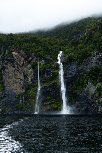 View of two waterfalls cascading down the rocky cliffs into the dark waters shrouded in mist a symphony of nature s power Milford Sound Southland Region New Zealand.