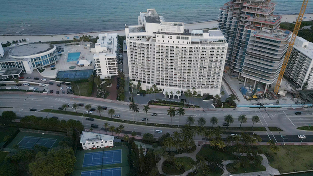 Aerial view of the Miami Beach coastline featuring a large white residential building a construction site tennis courts and palm trees in Miami Beach Florida United States. Aerial view of the Miami Beach coastline featuring a large white residential building a construction site tennis courts and palm trees in Miami Beach Florida United States.