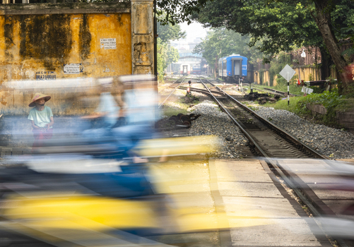 Hanoi Vietnam 11 November 2025 View of a person in a conical hat sitting by the train tracks in Hanoi with motion blur from a passing vehicle and a train in the distance.