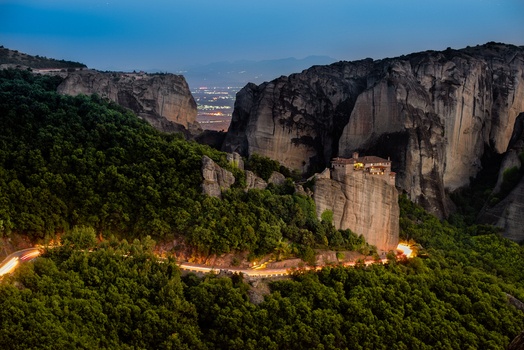 Aerial view of Meteora monastery perched on a sandstone rock formation surrounded by lush green forest and a winding road with light trails at dusk in Trikala Greece. Aerial view of Meteora monastery perched on a sandstone rock formation surrounded by lush green forest and a winding road with light trails at dusk in Trikala Greece.