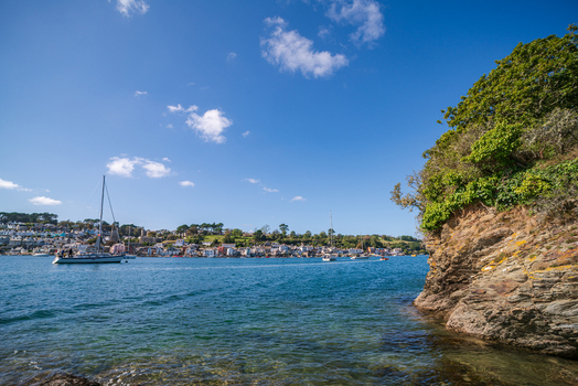 View of the Fowey River with sailboats moored near the historic town of Fowey featuring rocky cliffs lush greenery and a clear blue sky in Fowey Cornwall United Kingdom. View of the Fowey River with sailboats moored near the historic town of Fowey featuring rocky cliffs lush greenery and a clear blue sky in Fowey Cornwall United Kingdom.