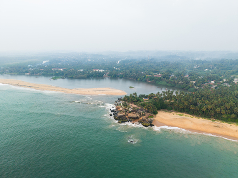 Aerial view of the scenic coastline where a river meets the ocean featuring a sandy beach rocky outcrop and dense palm trees under a hazy sky in Galle Southern Province Sri Lanka.