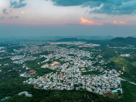 Aerial view of the city of Udaipur featuring dense residential architecture surrounding lush green hills and a distant lake under a twilight sky in Udaipur Rajasthan India.