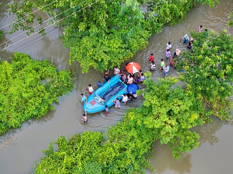 Banshkhali Upazila Bangladesh 10 July 2026 Aerial view of people wading through floodwaters and guiding a small blue boat through a submerged area surrounded by lush green trees. Banshkhali Upazila Bangladesh 10 July 2026 Aerial view of people wading through floodwaters and guiding a small blue boat through a submerged area surrounded by lush green trees.