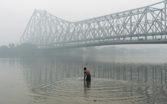 View of a man waist deep in the calm river water ripples expanding under the iconic Howrah Bridge shrouded in mist Kolkata West Bengal India. View of a man waist deep in the calm river water ripples expanding under the iconic Howrah Bridge shrouded in mist Kolkata West Bengal India.