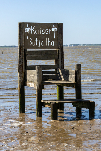 View of the Kaiser Butjatha wooden chair sculpture standing on the muddy shore of the Wadden Sea under a clear blue sky in Varel Lower Saxony Germany. View of the Kaiser Butjatha wooden chair sculpture standing on the muddy shore of the Wadden Sea under a clear blue sky in Varel Lower Saxony Germany.