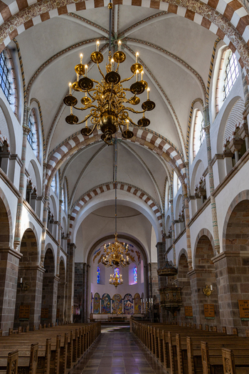 View of the interior of Ribe Cathedral featuring Romanesque arches vaulted ceilings and ornate golden chandeliers hanging above wooden pews in Ribe Denmark. View of the interior of Ribe Cathedral featuring Romanesque arches vaulted ceilings and ornate golden chandeliers hanging above wooden pews in Ribe Denmark.