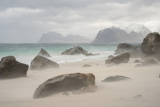 View of a sandy beach with large boulders and misty wind blowing sand set against a backdrop of snow capped mountains under an overcast sky in Leknes Lofoten Norway.