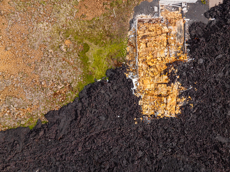 Aerial view of a house being consumed by a slow moving lava flow with black volcanic rock surrounding the wooden structure and green mossy terrain in Iceland. Aerial view of a house being consumed by a slow moving lava flow with black volcanic rock surrounding the wooden structure and green mossy terrain in Iceland.