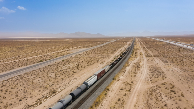 Aerial view of a long freight train traveling through the vast arid desert landscape with a parallel highway under a clear blue sky in the United States.