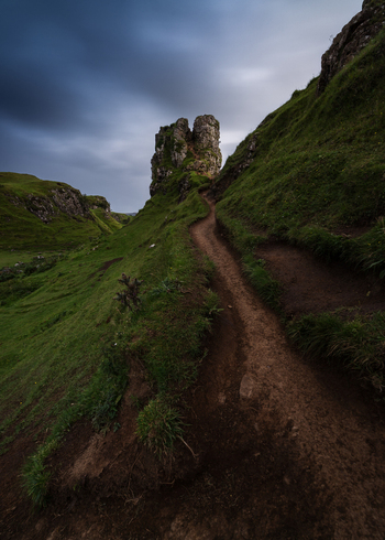 View of a narrow dirt path winding through the lush green hills of Fairy Glen towards the Castle Ewen rock formation under a dramatic cloudy sky in United Kingdom. View of a narrow dirt path winding through the lush green hills of Fairy Glen towards the Castle Ewen rock formation under a dramatic cloudy sky in United Kingdom.