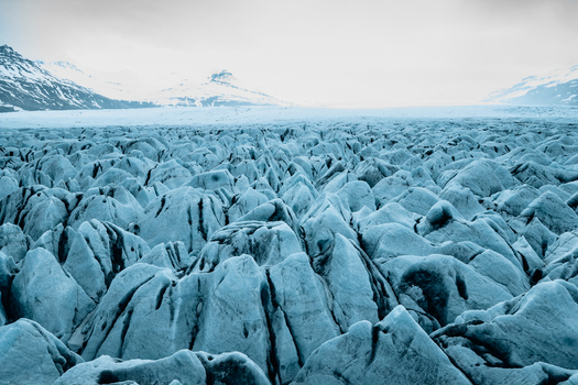 Aerial view of the jagged blue ice formations of Vatnajokull glacier with snow capped mountains in the background under an overcast sky in Iceland.