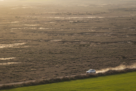 View of a white car driving on a dusty road through the arid landscape of Bardenas Reales with green fields in the foreground Navarre Navarre Spain. View of a white car driving on a dusty road through the arid landscape of Bardenas Reales with green fields in the foreground Navarre Navarre Spain.