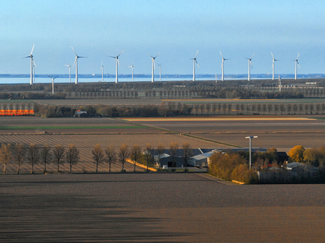 Aerial view of a row of modern wind turbines standing tall over vast agricultural fields and farm buildings under a clear blue sky in Netherlands.