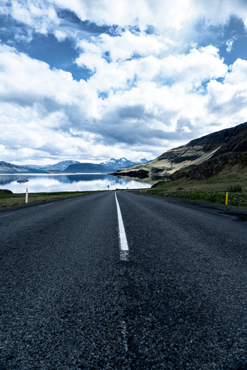 View of an asphalt road leading towards the calm waters of Hvalfjordur fjord under a cloudy sky with distant mountains in Iceland. View of an asphalt road leading towards the calm waters of Hvalfjordur fjord under a cloudy sky with distant mountains in Iceland.