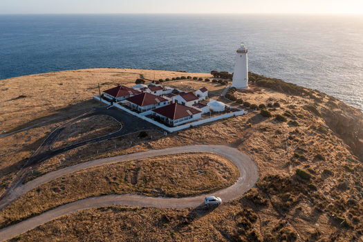 Aerial view of Cape Willoughby Lighthouse on a coastal cliff with historic buildings a winding road and the ocean under golden hour light in Kangaroo Island South Australia Australia.