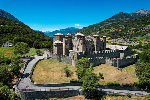 Aerial view of the medieval Fenis Castle with its stone towers and crenellated walls surrounded by lush green hills and alpine mountains in Fenis Aosta Valley Italy. Aerial view of the medieval Fenis Castle with its stone towers and crenellated walls surrounded by lush green hills and alpine mountains in Fenis Aosta Valley Italy.