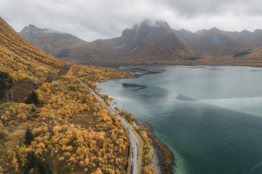 Aerial view of the Lofoten landscape featuring a winding coastal road vibrant autumn foliage on the hillsides and calm turquoise waters under a cloudy sky in Svolvaer Nordland Norway.