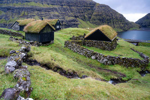 View of traditional black wooden houses with green turf roofs and stone walls nestled in a lush green valley with mountains and a stream in the Faroe Islands.