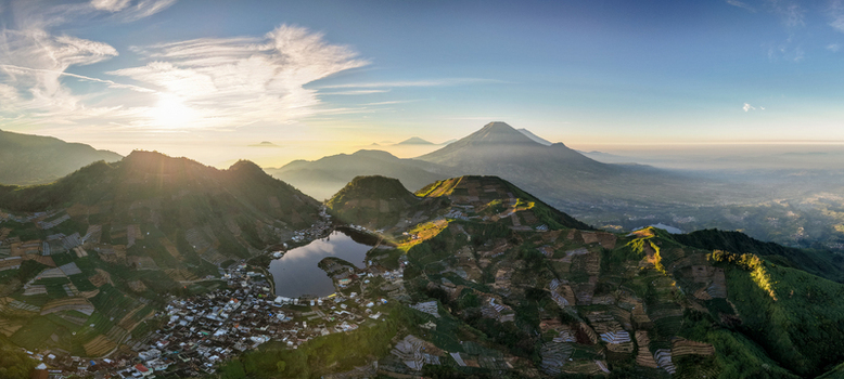 Aerial view of Telaga Cebong lake surrounded by terraced fields and Sikunang village at sunrise with distant volcanoes in Kecamatan Kejajar Jawa Tengah Indonesia.