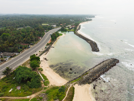 Aerial view of the tropical coastline featuring a stone breakwater sandy beach and a coastal road lined with palm trees in Koggala Southern Province Sri Lanka. Aerial view of the tropical coastline featuring a stone breakwater sandy beach and a coastal road lined with palm trees in Koggala Southern Province Sri Lanka.