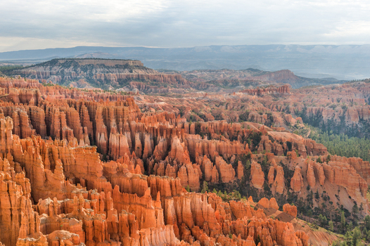 View of the iconic orange hoodoos and rock formations of Bryce Canyon National Park with pine trees and distant mountains under a cloudy sky in Utah United States.