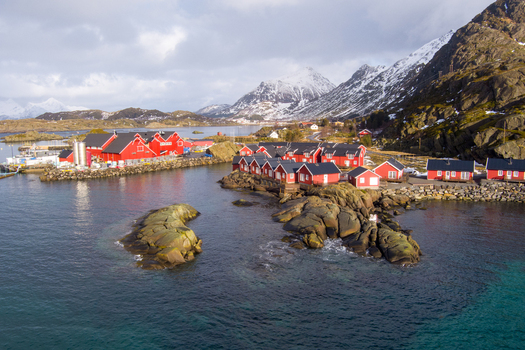 Aerial view of the iconic red rorbuer cabins in Lofoten nestled along the rocky coastline with snow capped mountains in the background Nordland Norway.