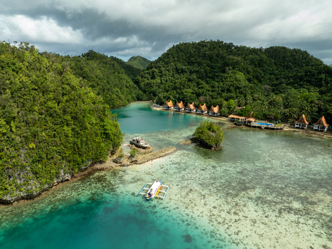Aerial view of Bucas Grande featuring lush green hills turquoise waters a traditional outrigger boat and small wooden cottages along the shore in Siargao Philippines. Aerial view of Bucas Grande featuring lush green hills turquoise waters a traditional outrigger boat and small wooden cottages along the shore in Siargao Philippines.