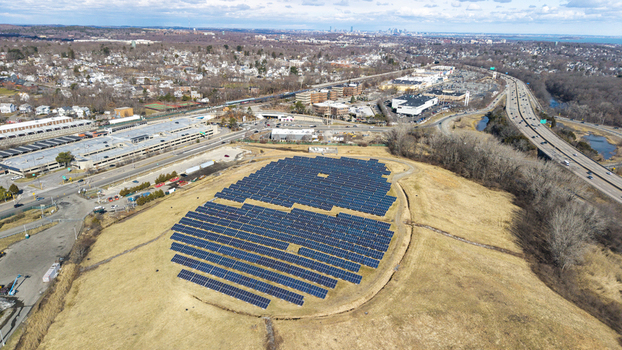 Aerial view of rows of solar panels glinting under the sun on a grassy hill near a highway contrasting with the urban sprawl Braintree Massachusetts United States.