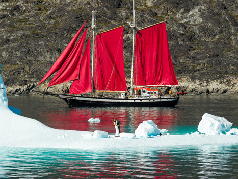 Aerial view of a couple standing on a large iceberg near a classic sailboat with vibrant red sails in the icy waters of Greenland. Aerial view of a couple standing on a large iceberg near a classic sailboat with vibrant red sails in the icy waters of Greenland.