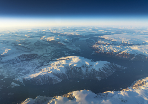 Aerial view of snow covered mountain peaks and icy fjords under a clear blue sky at the horizon in Greenland. Aerial view of snow covered mountain peaks and icy fjords under a clear blue sky at the horizon in Greenland.