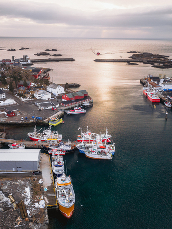 Aerial view of boats nestled in the harbor vibrant against the deep blue water with red buildings dotting the shoreline Ballstad Nordland Norway. Aerial view of boats nestled in the harbor vibrant against the deep blue water with red buildings dotting the shoreline Ballstad Nordland Norway.