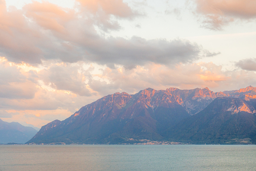 View of Lake Geneva with the Alps in the background during golden hour featuring soft pink and orange clouds over the mountains in Bourg en Lavaux Vaud Switzerland. View of Lake Geneva with the Alps in the background during golden hour featuring soft pink and orange clouds over the mountains in Bourg en Lavaux Vaud Switzerland.