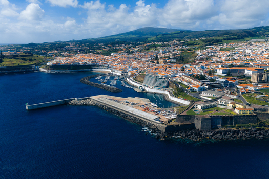 Aerial view of the historic coastal city featuring white buildings with terracotta roofs a harbor with breakwaters and lush green hills under a blue sky Angra do Heroismo Azores Portugal.
