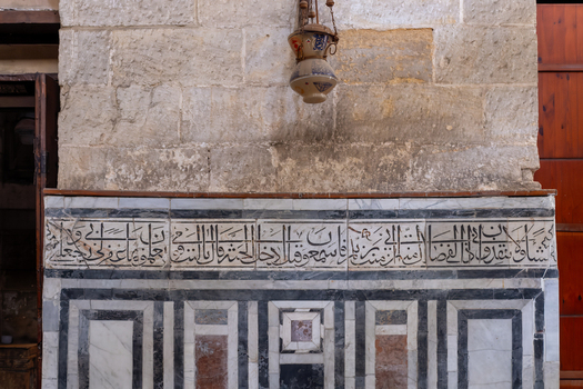 View of the Sultan al Ghuri Complex featuring intricate Arabic calligraphy and marble inlay on a stone wall with a hanging lamp in El Darb El Ahmar Cairo Governorate Egypt. View of the Sultan al Ghuri Complex featuring intricate Arabic calligraphy and marble inlay on a stone wall with a hanging lamp in El Darb El Ahmar Cairo Governorate Egypt.
