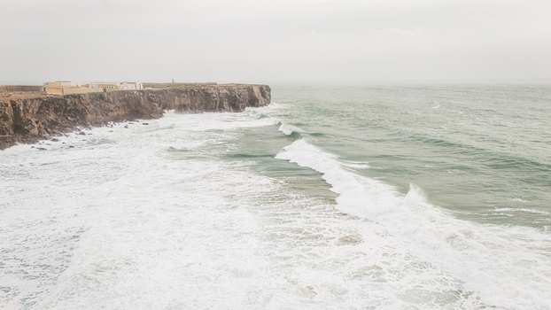 Aerial view of the Sagres Fortress on rugged cliffs with crashing waves and white foam under a misty sky in Sagres Faro Portugal. Aerial view of the Sagres Fortress on rugged cliffs with crashing waves and white foam under a misty sky in Sagres Faro Portugal.