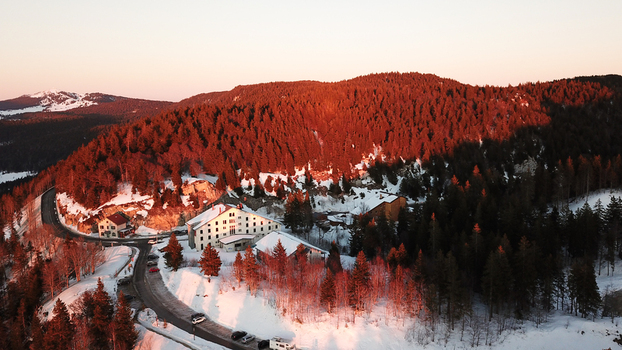 Aerial view of Col de la Faucille featuring snow covered buildings a winding road and dense pine forests illuminated by golden hour light in Gex Auvergne Rhone Alpes France.