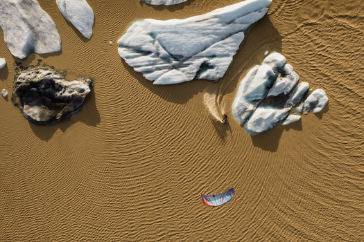 Aerial view of a kite surfer gliding across the sandy shore near Vatnajokull with scattered icebergs resting on the beach in Iceland.