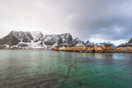 View of quaint yellow wooden houses nestling along the tranquil turquoise waters against a backdrop of snow dusted mountains Hamn&oslash;y Nordland Norway.
