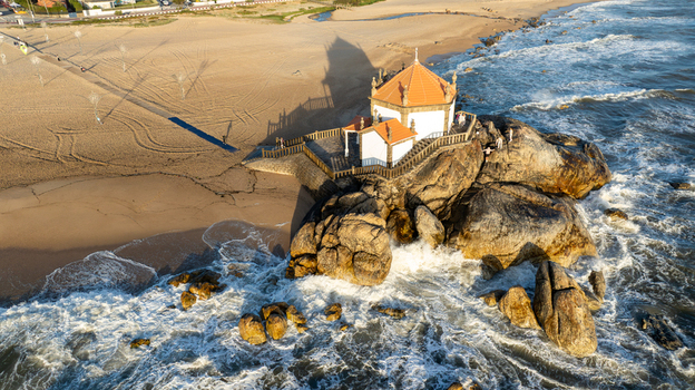 Aerial view of Capela do Senhor da Pedra on a rocky outcrop with waves crashing and long shadows on the sandy beach in Miramar Porto Portugal.