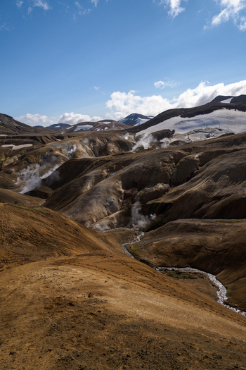View of a landscape dominated by geothermal activity with steaming vents and snowy mountains under a bright blue sky in Hrunamannahreppur Hrunamannahreppur Iceland.